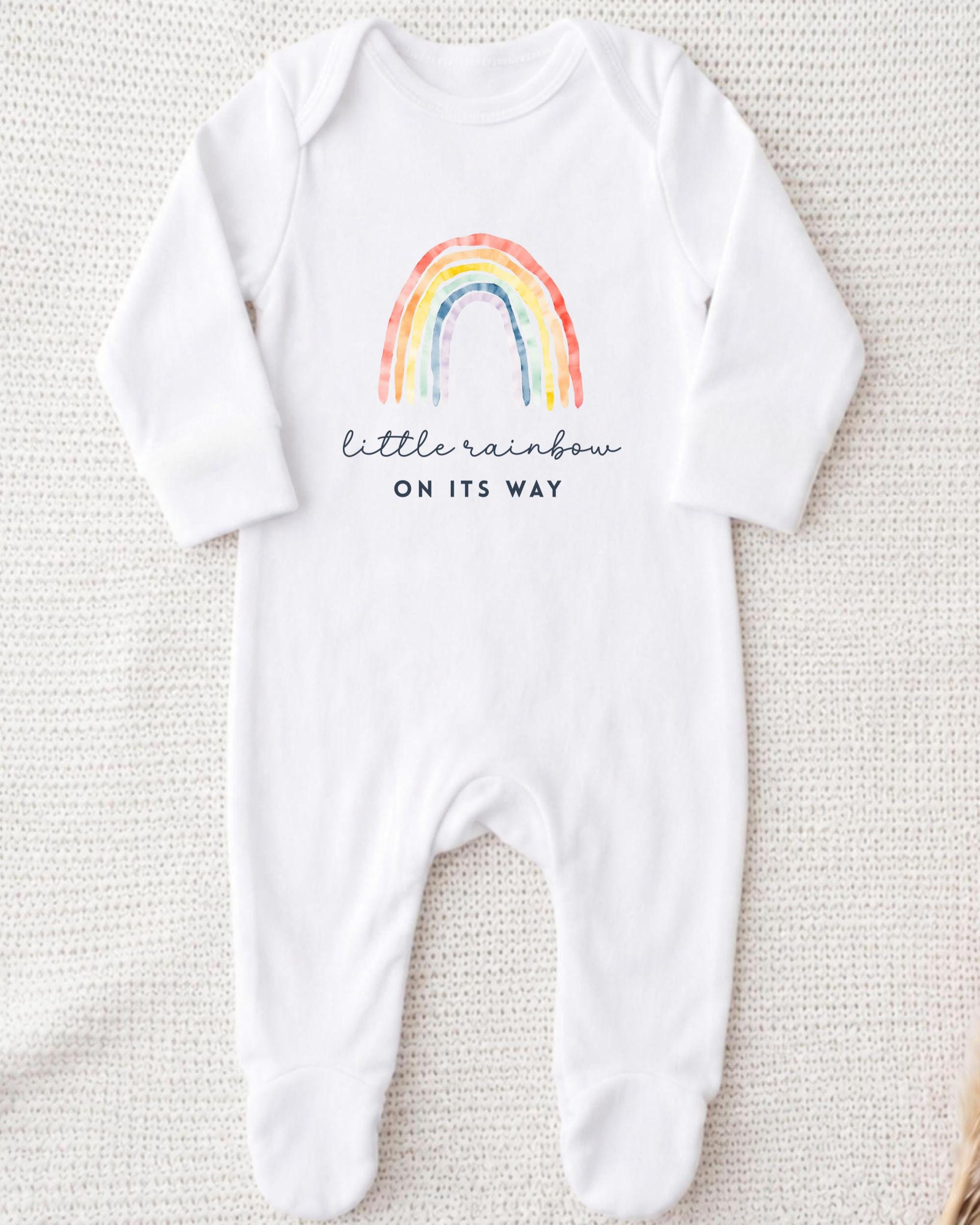 A white onesie with a rainbow graphic and the text "little rainbow on its way" printed on it.