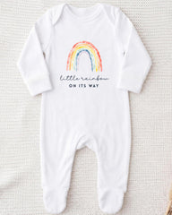 A white onesie with a rainbow graphic and the text "little rainbow on its way" printed on it.
