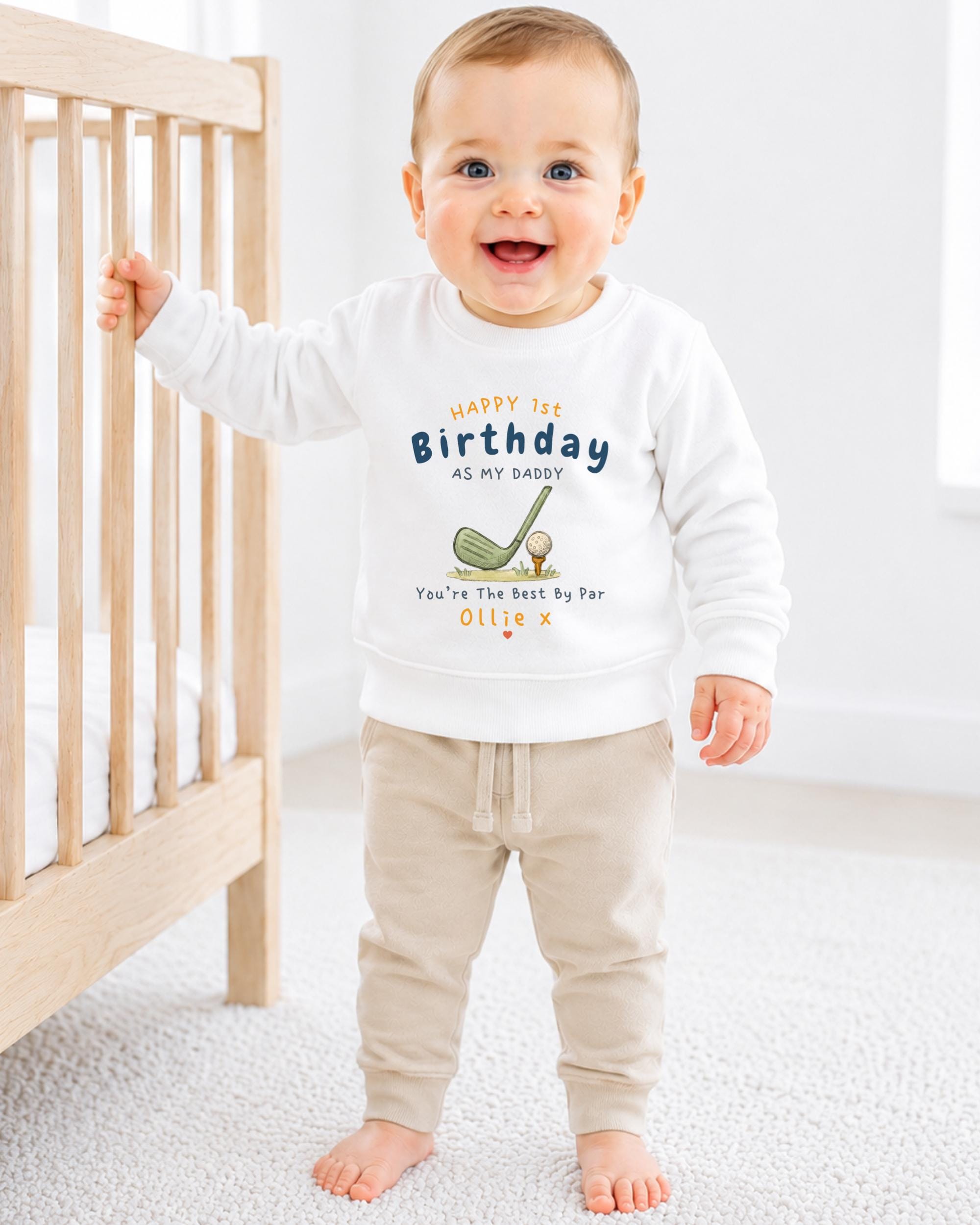 A smiling baby boy wearing a white sweatshirt with a birthday message and a golf club graphic, standing next to a wooden crib.