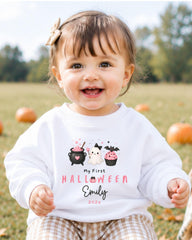 A smiling baby girl wearing a white sweater with a Halloween-themed design and text.