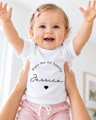 A smiling baby girl wearing a white t-shirt with the text "Pass me to Aunt Jessica" and pink shorts, being held up by someone's arms.
