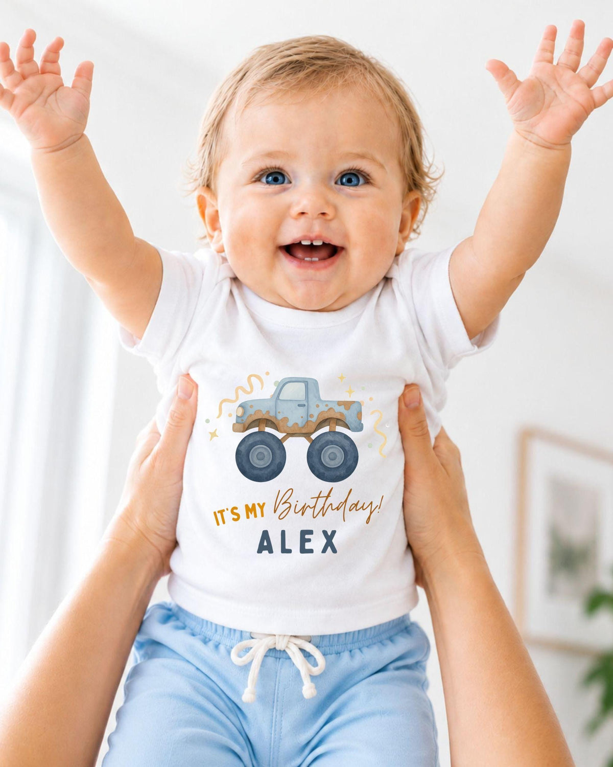 A smiling baby wearing a white t-shirt with a truck graphic and the text "It's my birthday Alex" is being held up by someone's arms.