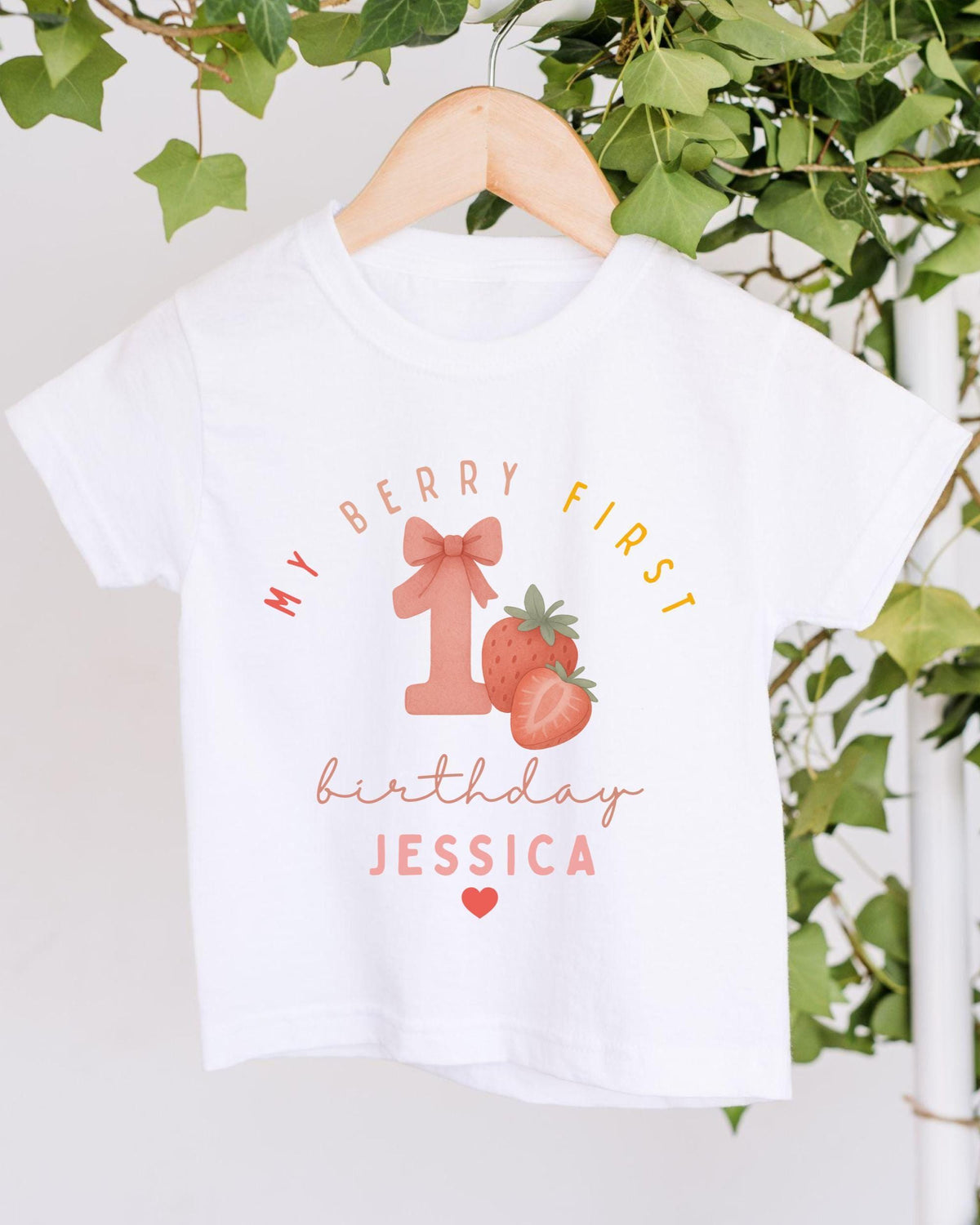 A white t-shirt with a graphic design featuring a pink number one, strawberries, and the text "My Berry First birthday Jessica" printed on it.