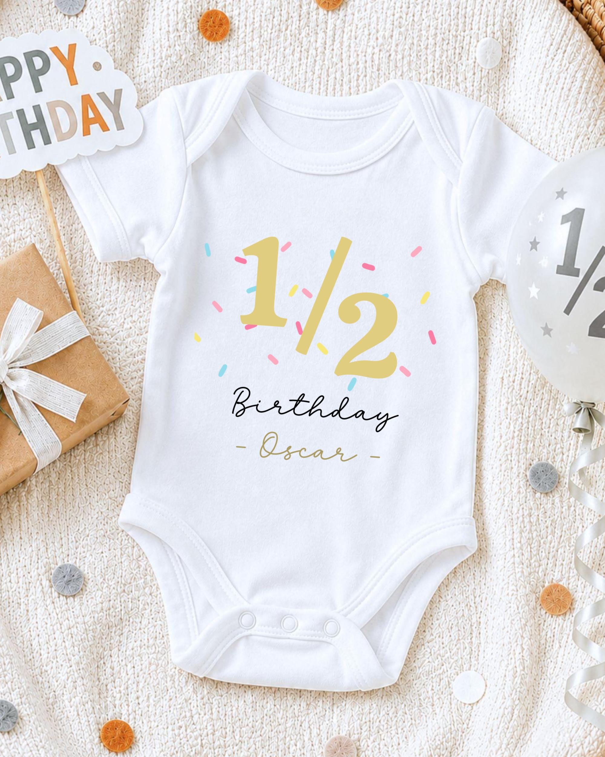 A white onesie with the text "1/2 Birthday Oscar" printed on it, along with a birthday sign and confetti, is displayed on a bed.