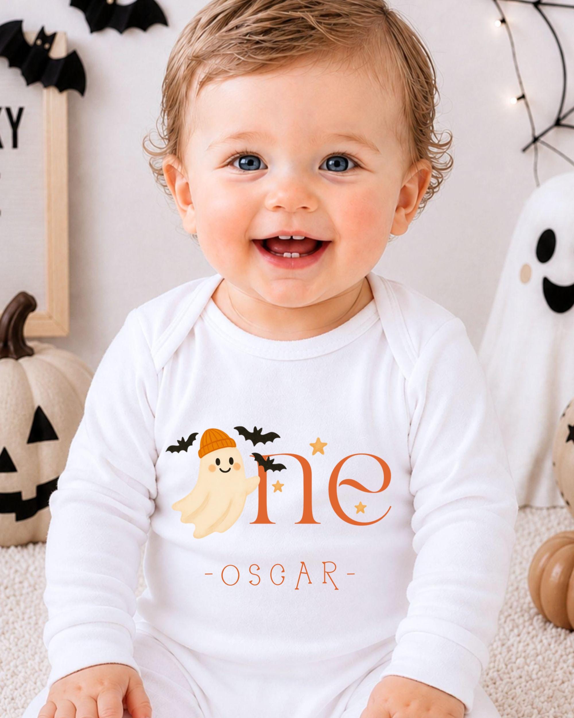 A smiling baby boy wearing a white onesie with a ghost and the text "One Oscar" is sitting in front of Halloween decorations.