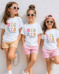 Three young girls wearing white t-shirts with colorful text and sunglasses, standing against a white brick wall.