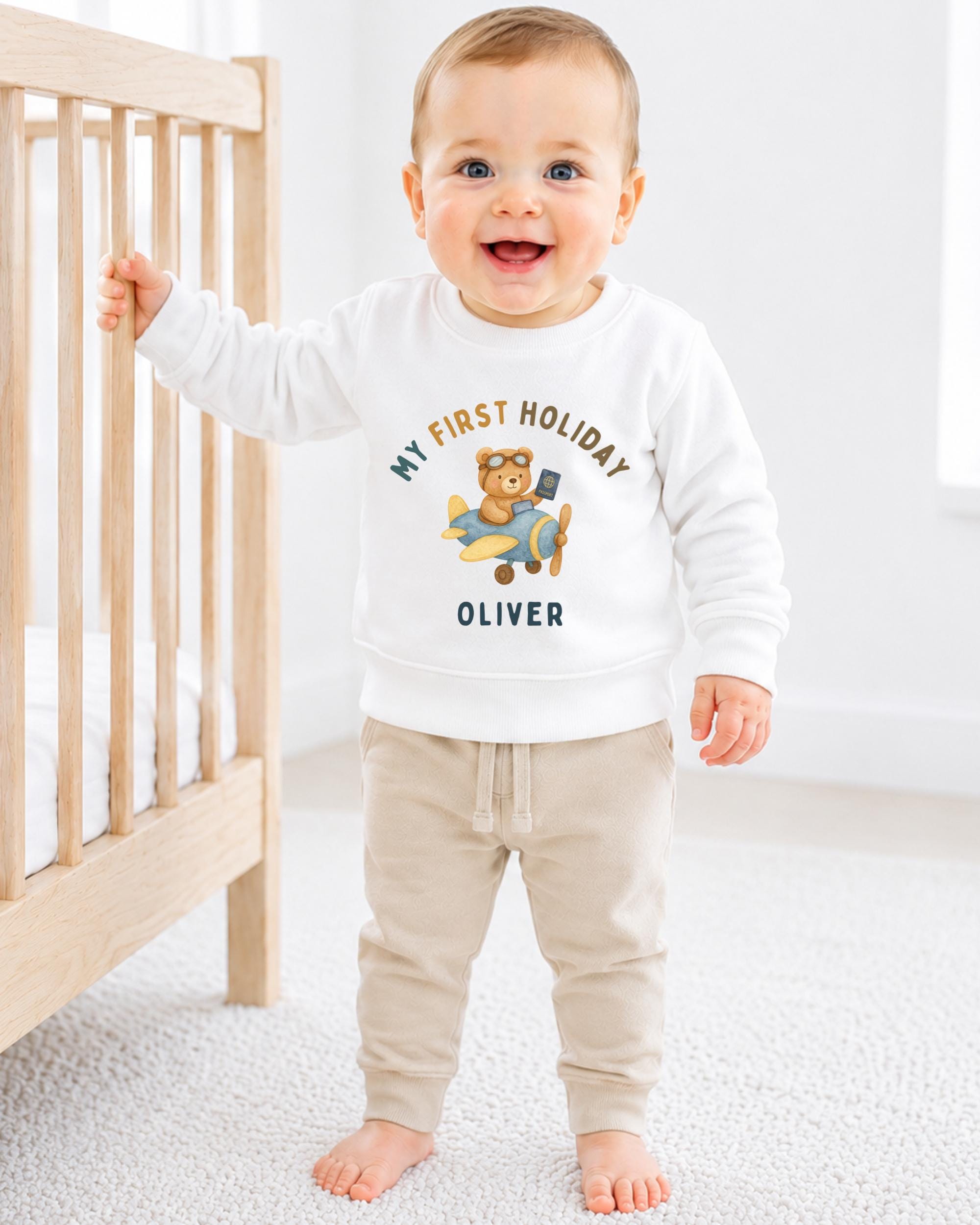A smiling baby boy wearing a white sweatshirt with a cartoon image of a bear and the text "My first holiday Oliver" stands next to a wooden crib.