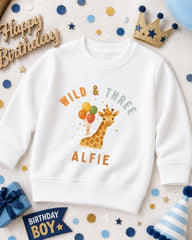A white sweatshirt with a giraffe design and the text "Wild & Three Alfie" printed on it, surrounded by birthday decorations such as a crown, confetti, and a birthday hat.
