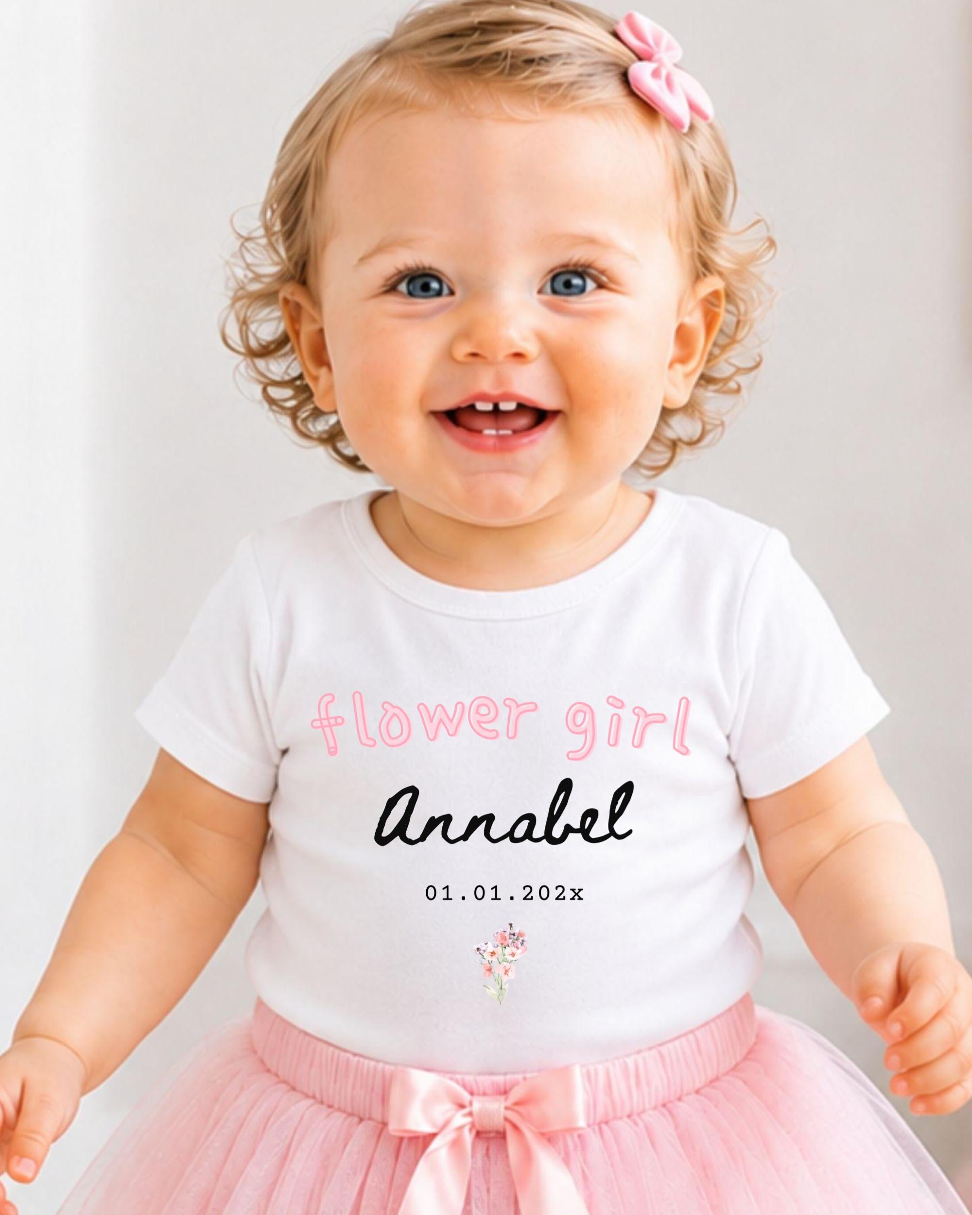 A smiling baby girl wearing a white t-shirt with the text "flower girl Annabel" and a pink tutu skirt.