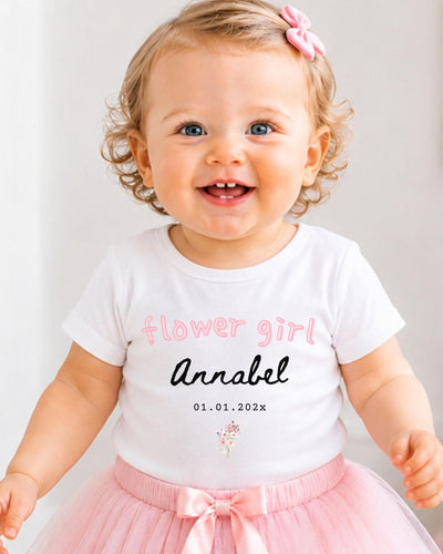 A smiling baby girl wearing a white t-shirt with the text "flower girl Annabel" and a pink tutu skirt.