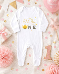 A white onesie with a bee design, surrounded by pink and gold decorations, including a pink balloon, a pink flower, and confetti.