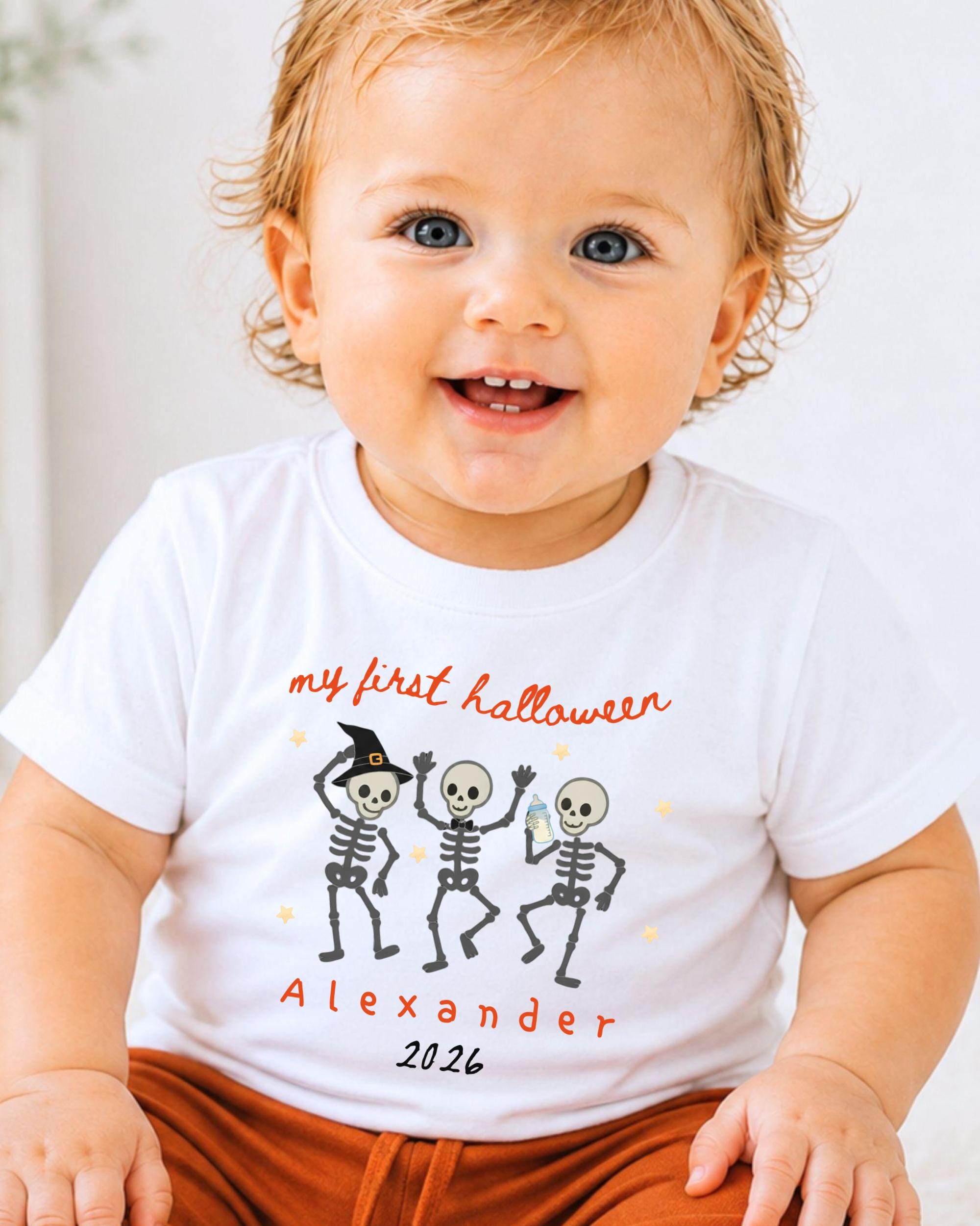 A smiling baby wearing a white t-shirt with a Halloween-themed design featuring three cartoon skeletons dancing.