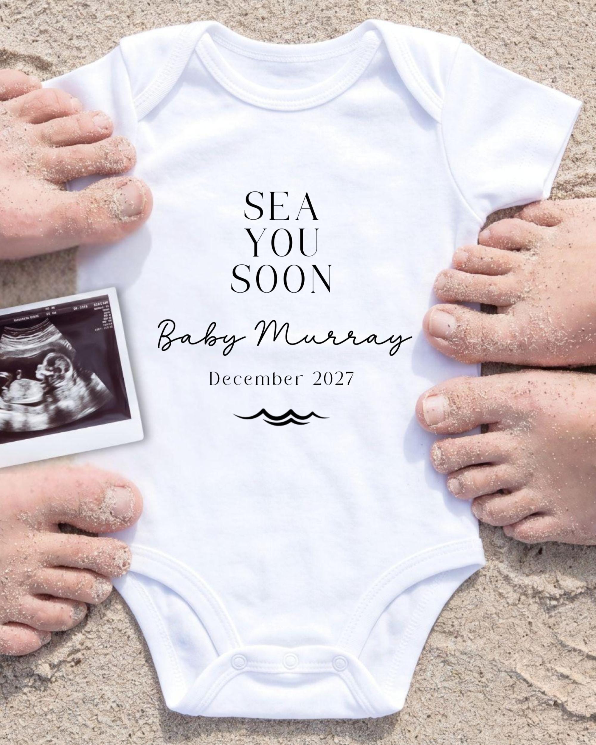 a white onesie with the text "SEA YOU SOON Baby Murray December 2027" printed on it, surrounded by sand and a black and white photograph of a baby's belly.