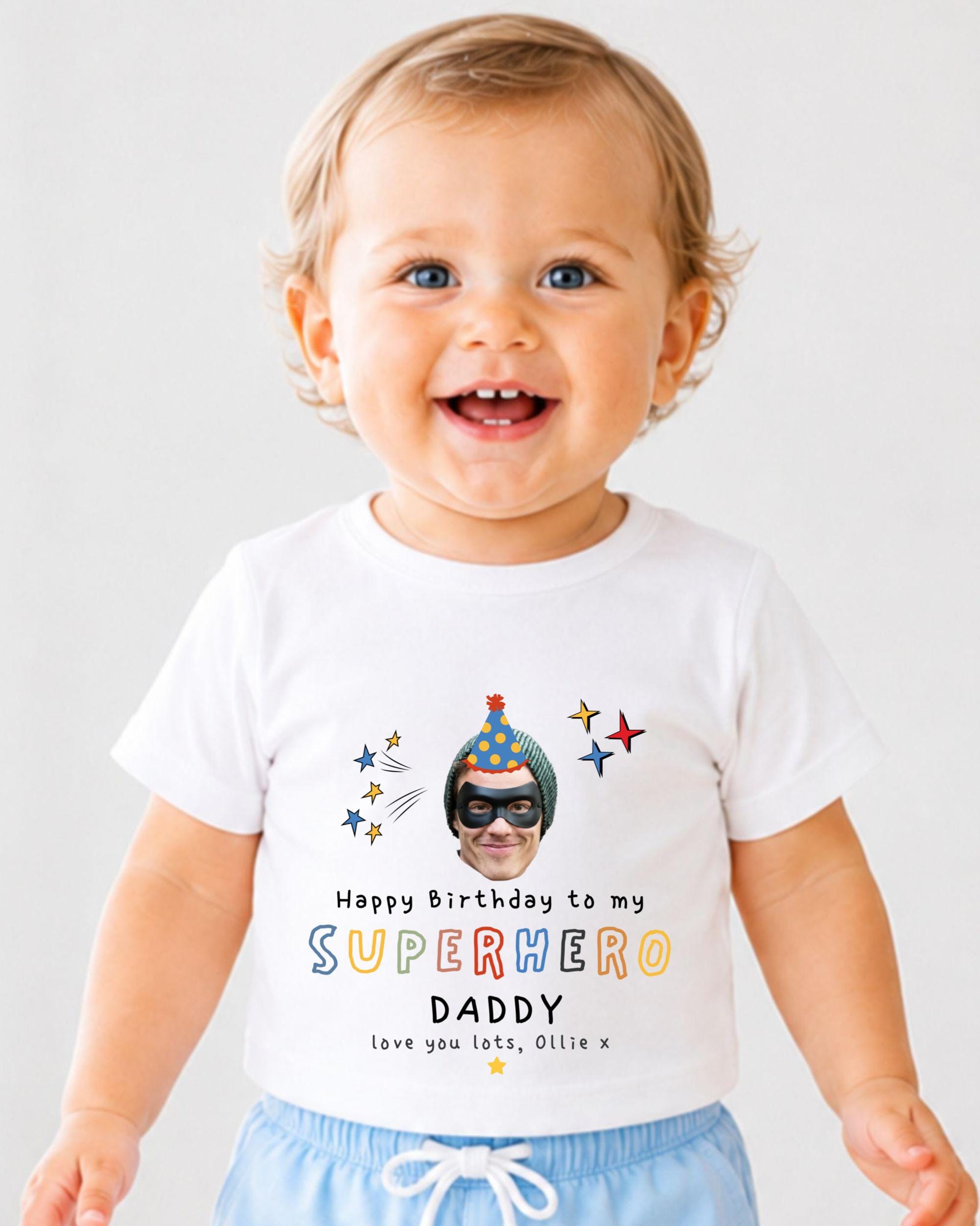 A smiling baby wearing a white t-shirt with a personalized birthday message and a superhero image printed on it.