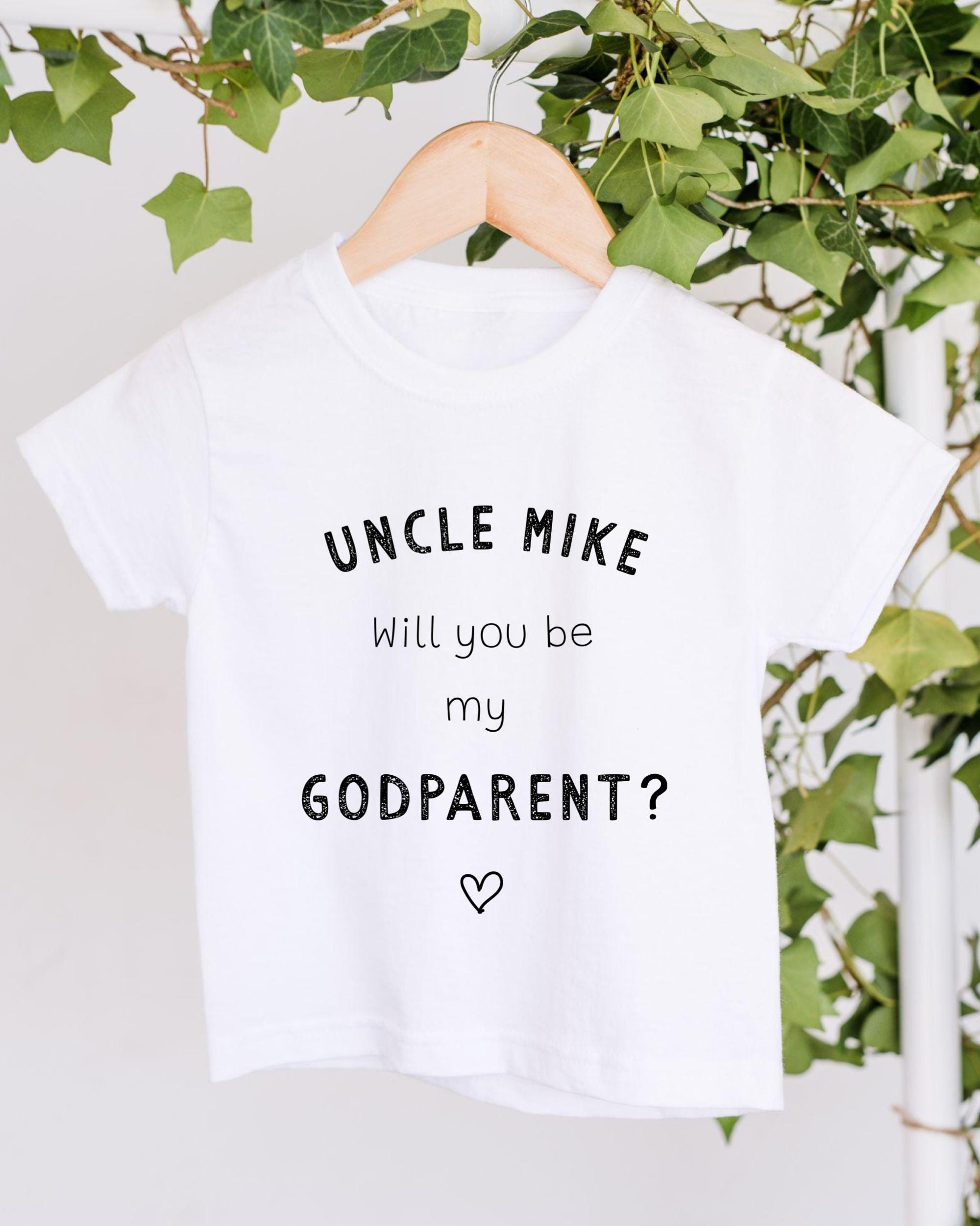 A white t-shirt with the text "UNCLE MIKE will you be my GODPARENT?" printed on it, hanging on a wooden hanger.