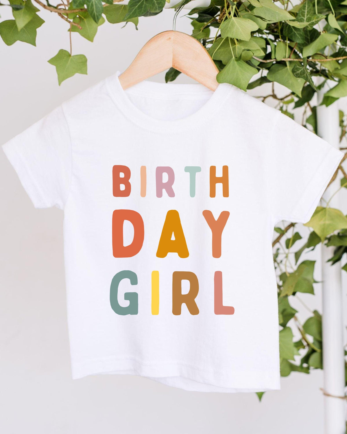 A white t-shirt with the text "BIRTHDAY GIRL" printed on it, hanging on a wooden hanger.