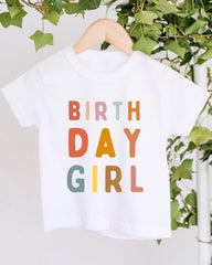 A white t-shirt with the text "BIRTHDAY GIRL" printed on it, hanging on a wooden hanger.