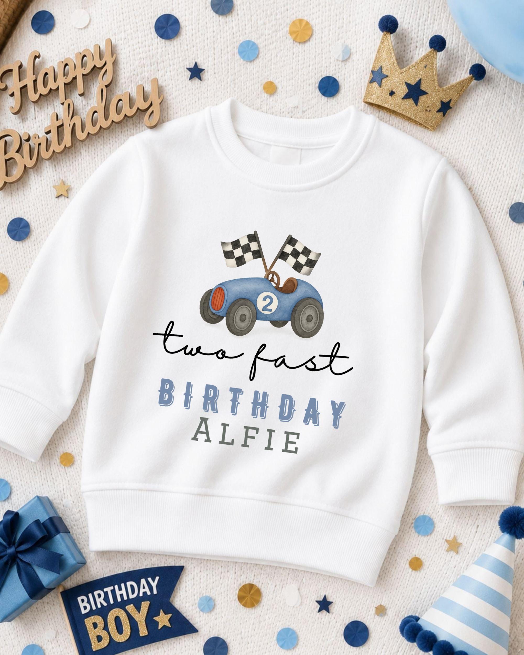 A white sweatshirt with a racing car design and the text "two fast birthday alfie" printed on it, surrounded by birthday decorations and a crown.