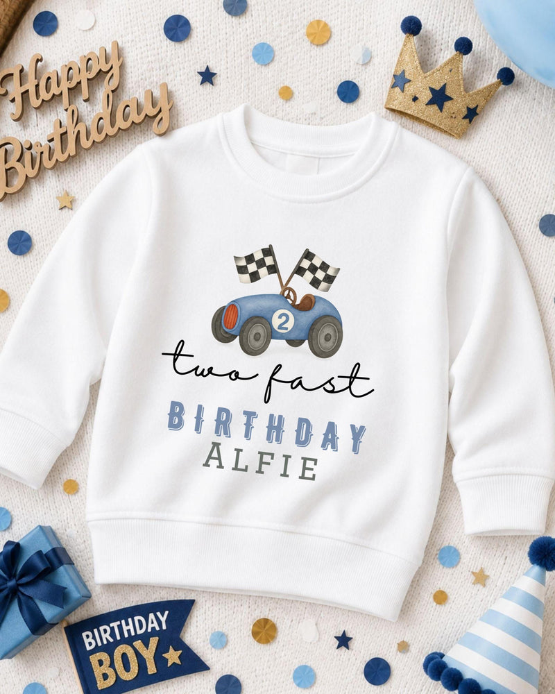 A white sweatshirt with a racing car design and the text "two fast birthday alfie" printed on it, surrounded by birthday decorations and a crown.