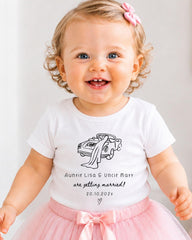 A smiling baby girl wearing a white t-shirt with a graphic of a car and text that says "Auntie Lisa & Uncle Matt are getting married! 20.10.2021".