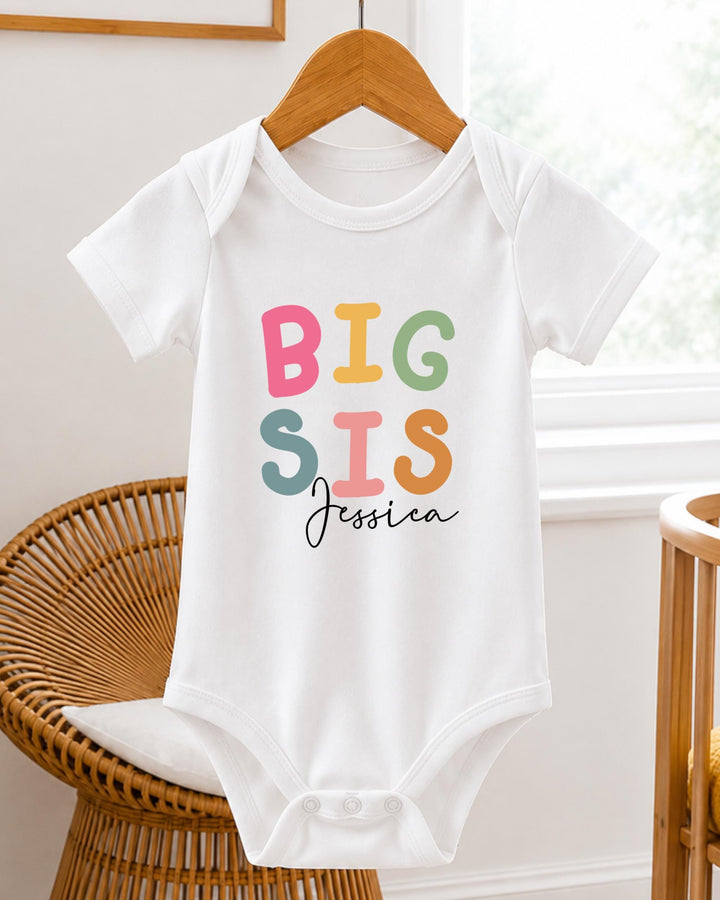 A white baby bodysuit with the text "BIG SIS Jessica" printed on it, hanging on a wooden hanger.