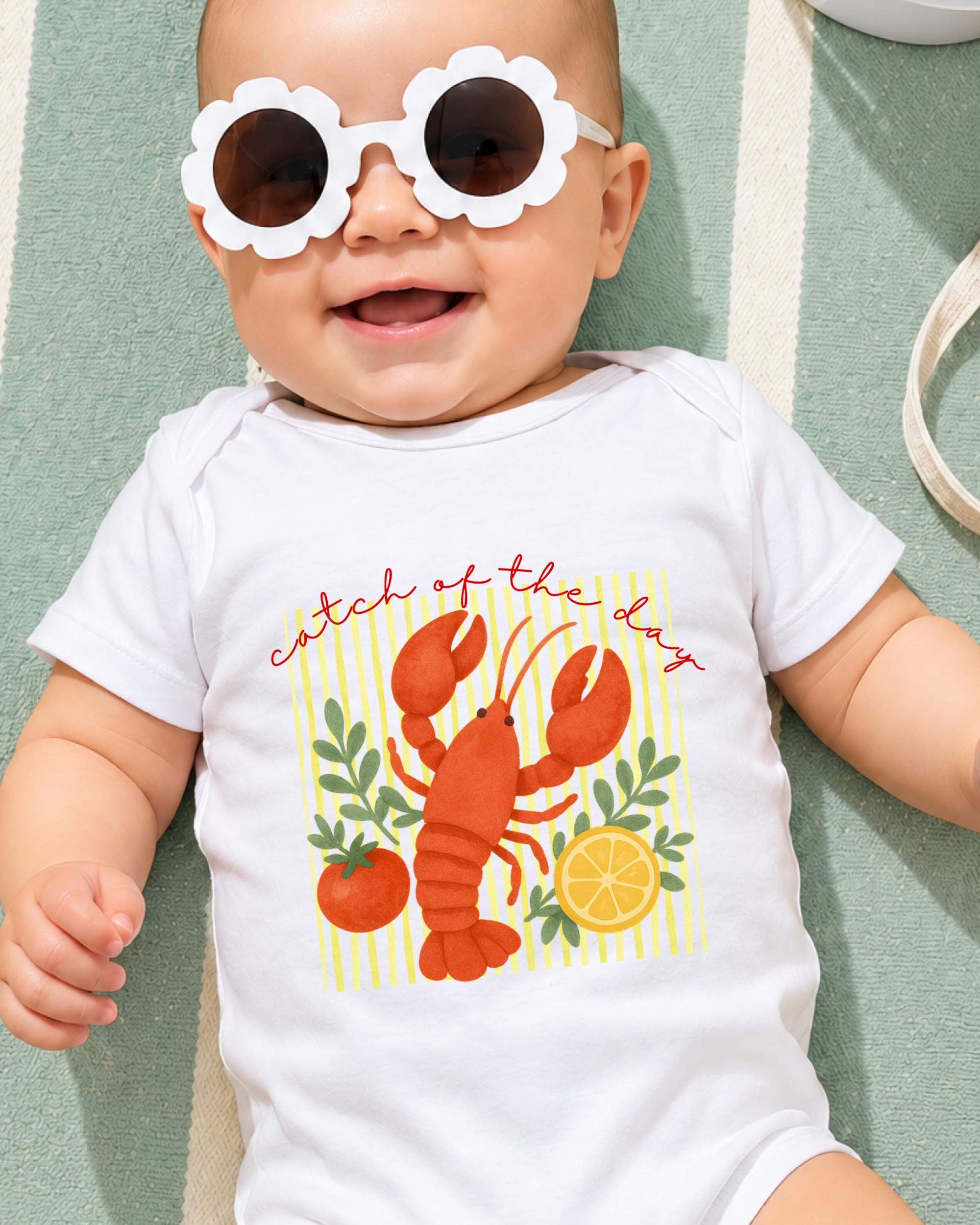 A baby wearing a white onesie with a lobster design and sunglasses lying on a green and white striped surface.
