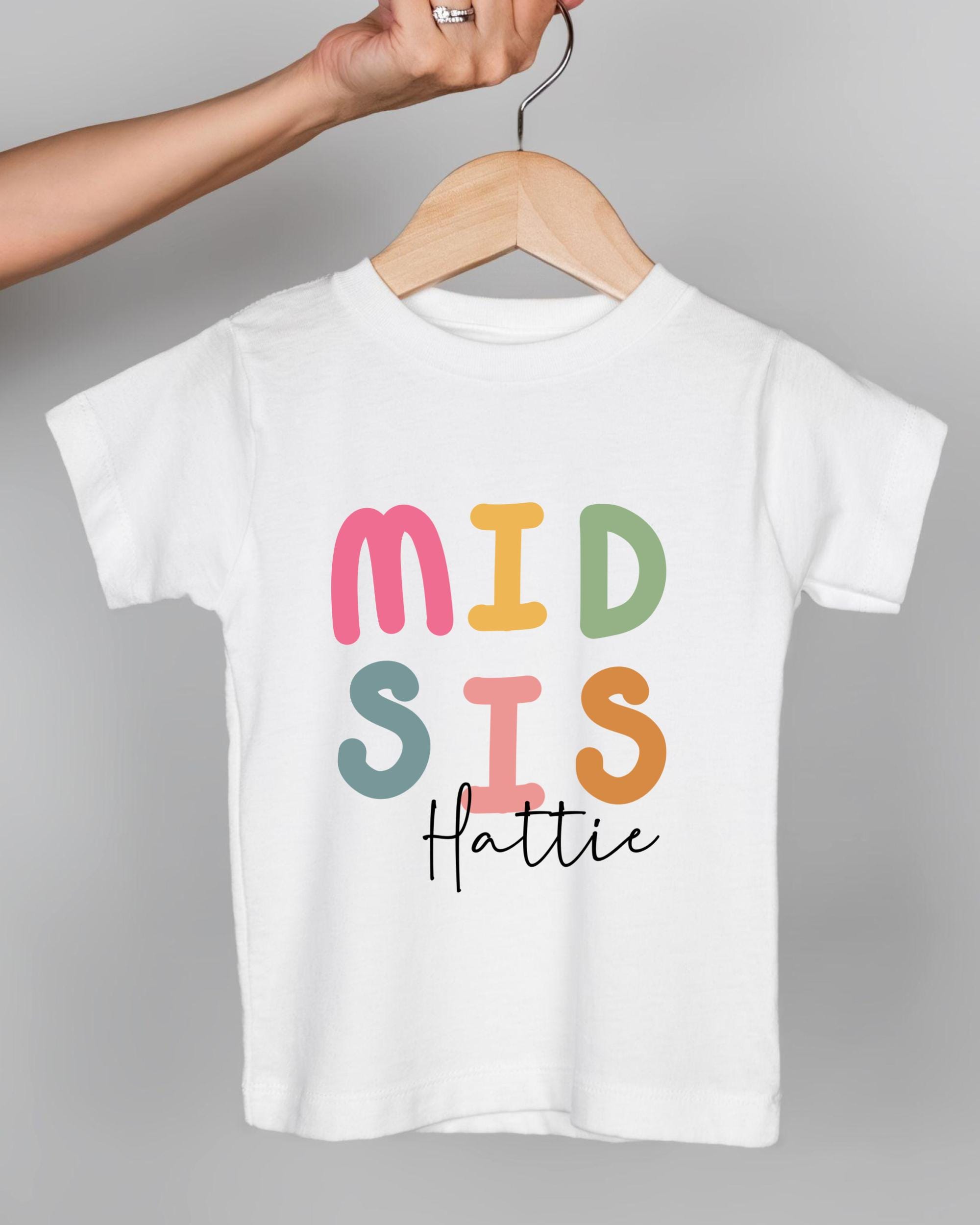 A person's hand is holding a white t-shirt with the text "MID SIS Hattie" printed on it.