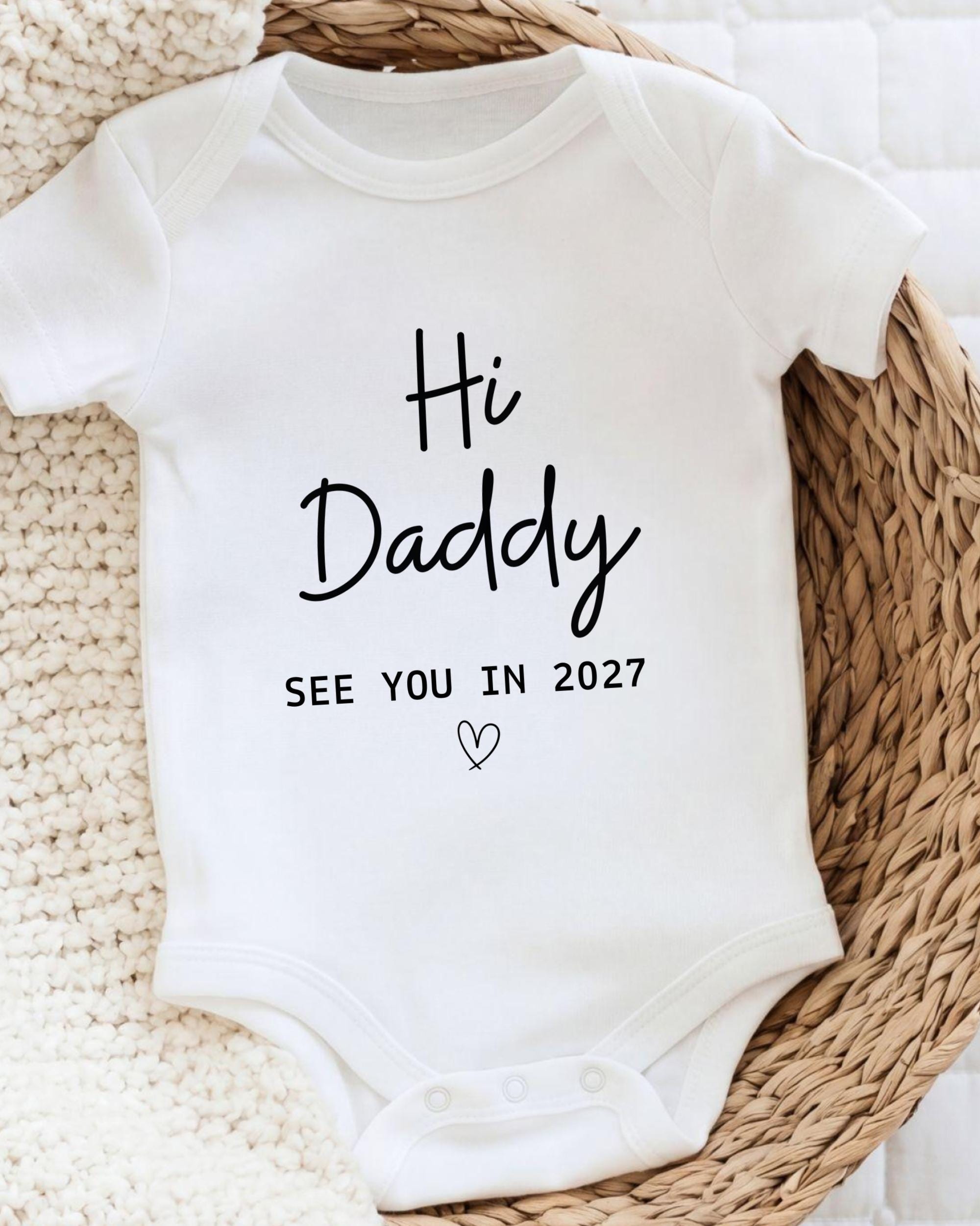 A white baby onesie with the text "Hi Daddy See You in 2027" printed on it.