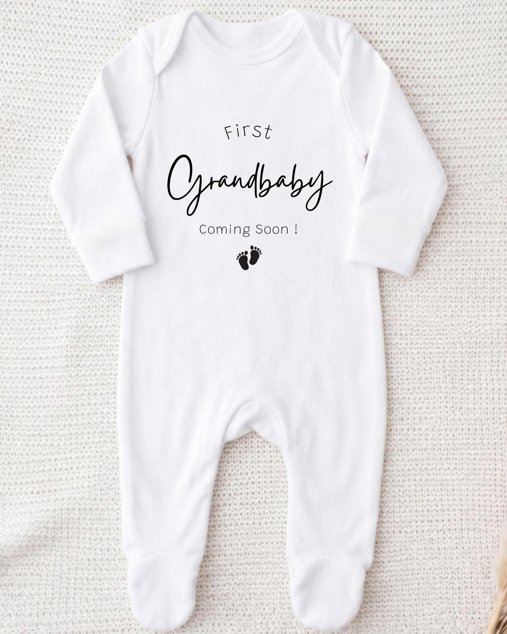 A white onesie with the text "First Grandbaby Coming Soon!" printed on it, along with a small black footprint icon.