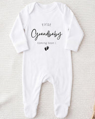 A white onesie with the text "First Grandbaby Coming Soon!" printed on it, along with a small black footprint icon.