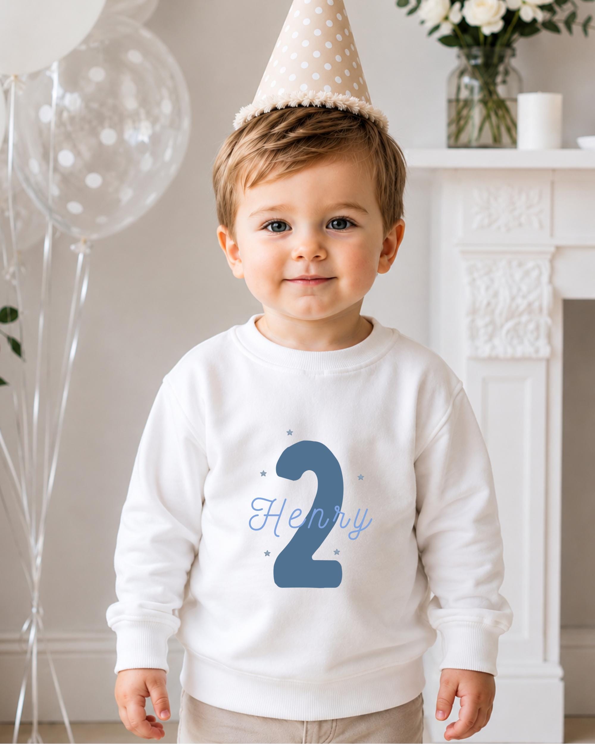 A young boy wearing a white sweatshirt with the number 2 and a birthday hat, standing in front of a fireplace with balloons and flowers.