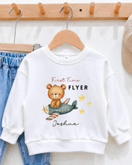 A white sweatshirt with a cartoon image of a bear riding an airplane, along with the text "First Time Flyer Joshua" printed on it.
