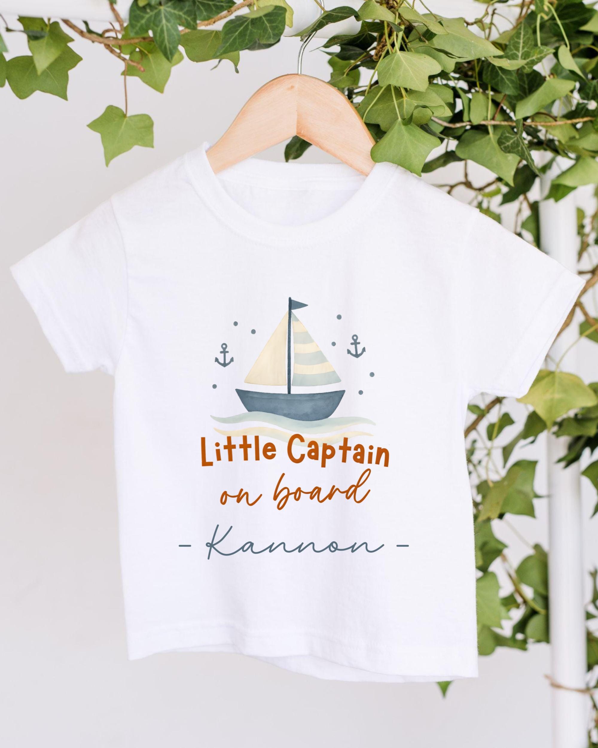 A white t-shirt with a sailboat graphic and the text "Little Captain on board" printed on it, hanging on a wooden hanger.