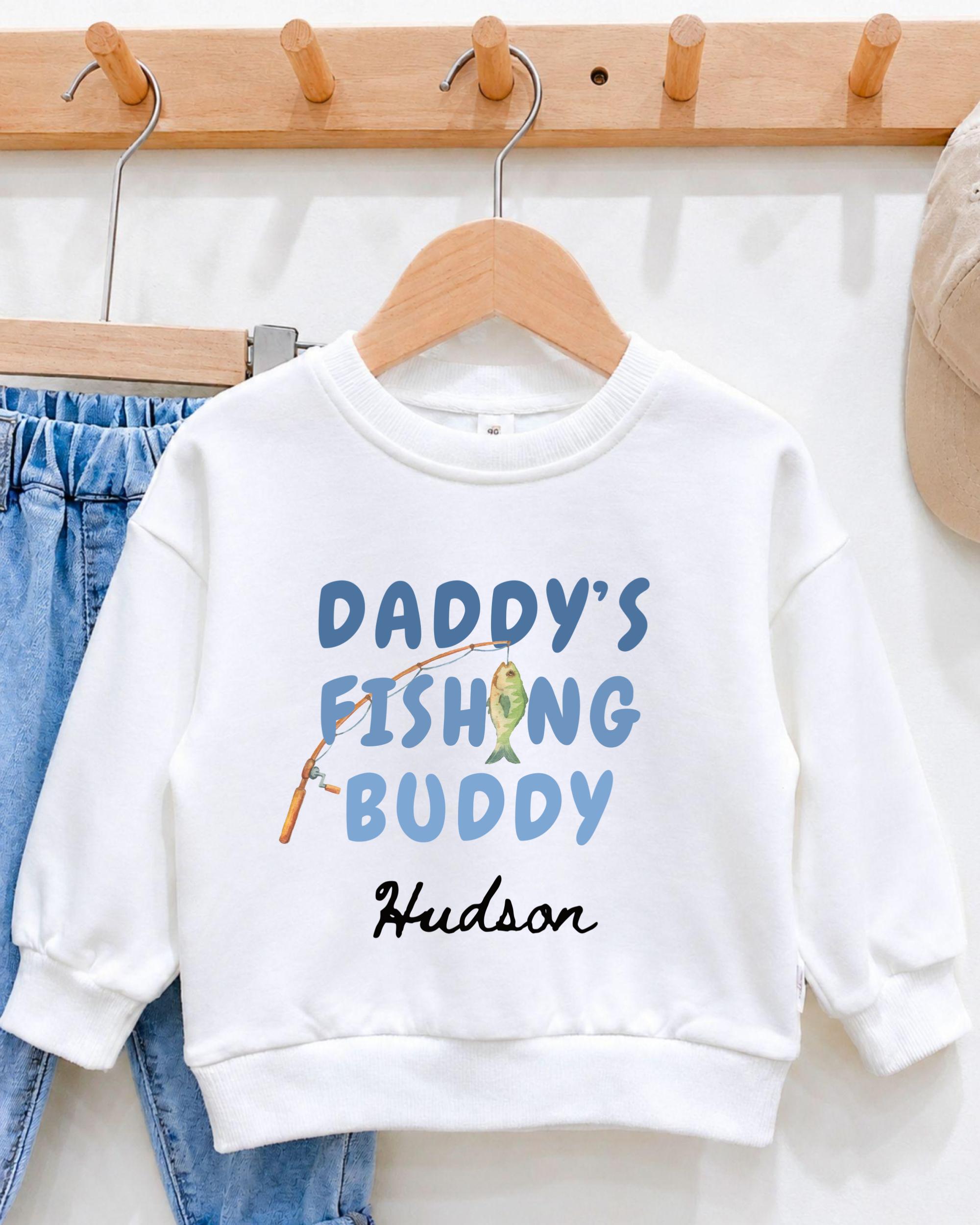 A white sweatshirt with the text "DADDY'S FISHING BUDDY" printed on it, along with a small illustration of a fish.