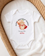 A white onesie with a graphic of a bear and the text "Happy Birthday Daddy" printed on it, placed on a white blanket.