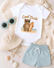 A baby's outfit, including a white onesie with a cartoon bear and the text "Cool Dude", a beige bucket hat, and light blue shorts, along with sunglasses and seashells, are arranged on a beige surface.