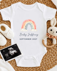 A white baby onesie with a rainbow graphic and the text "Baby Jeffrey" printed on it, along with a pair of white crocheted shoes and a rattle. There is also an " Ultrasound" image displayed, suggesting the baby's gender.