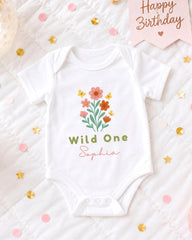 A white onesie with a flower design and the text "Wild One Sophia" printed on it, placed on a bed with confetti and a birthday card.
