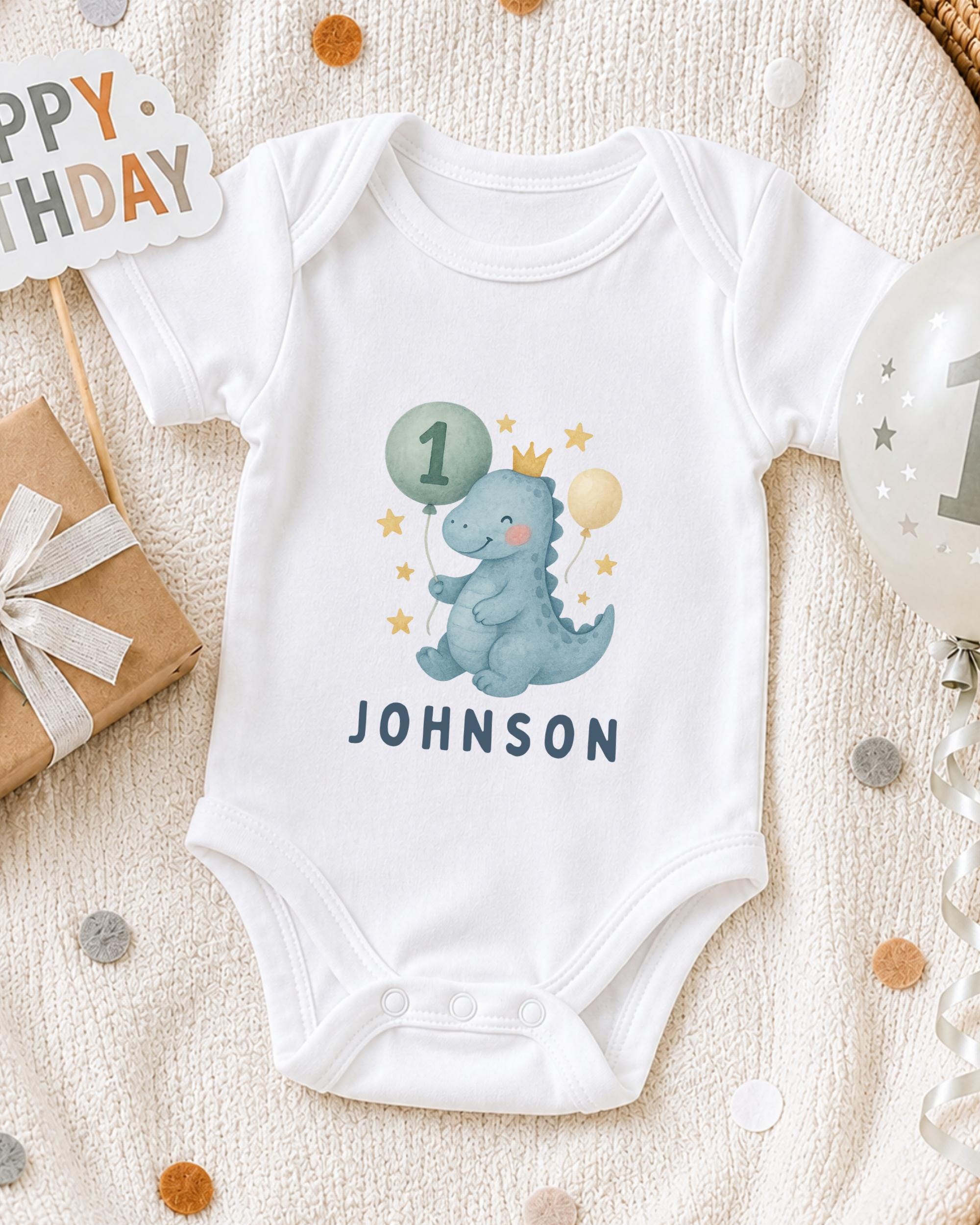 A white onesie with a dinosaur and the number 1, along with a "Happy Birthday" sign, is displayed on a bed with confetti.