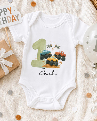 A white bodysuit with a graphic of a truck on it, along with a birthday sign and a balloon, all placed on a white blanket.
