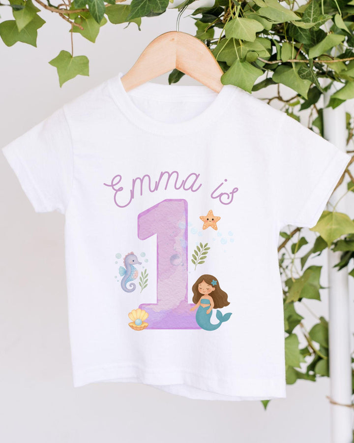 A white t-shirt with a mermaid and a number 1 printed on it, along with some marine-themed elements.
