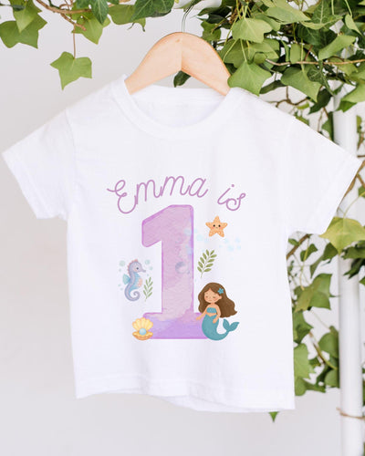 A white t-shirt with a mermaid and a number 1 printed on it, along with some marine-themed elements.