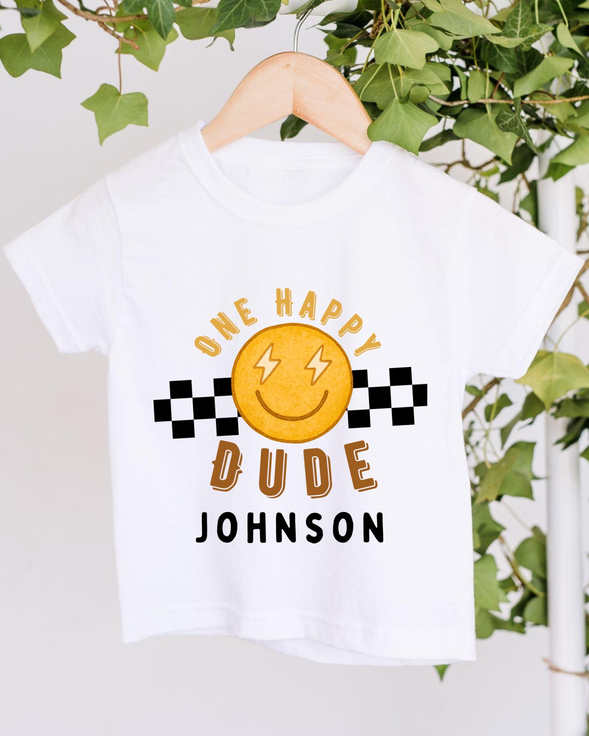 A white t-shirt with a cheerful smiley face and text printed on it, hanging on a wooden hanger.