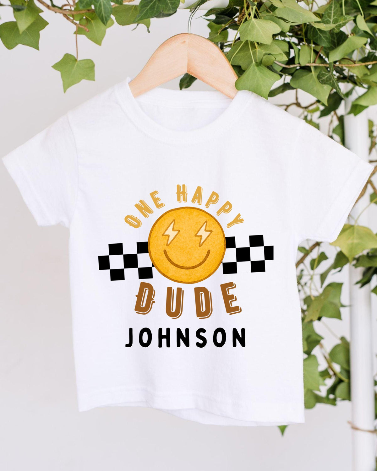 A white t-shirt with a cheerful smiley face and text printed on it, hanging on a wooden hanger.