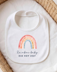 A white bib with a colorful rainbow graphic and the text "Rainbow baby due novely 2027" printed on it.