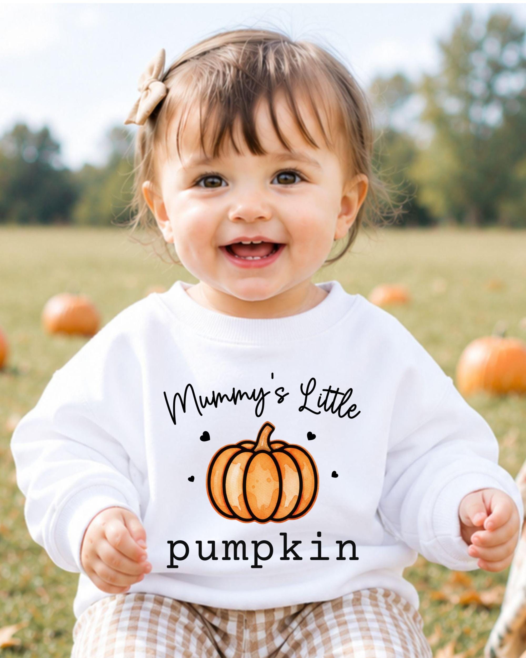A smiling baby girl wearing a white sweatshirt with a pumpkin graphic and the text "Mummy's Little pumpkin" printed on it.