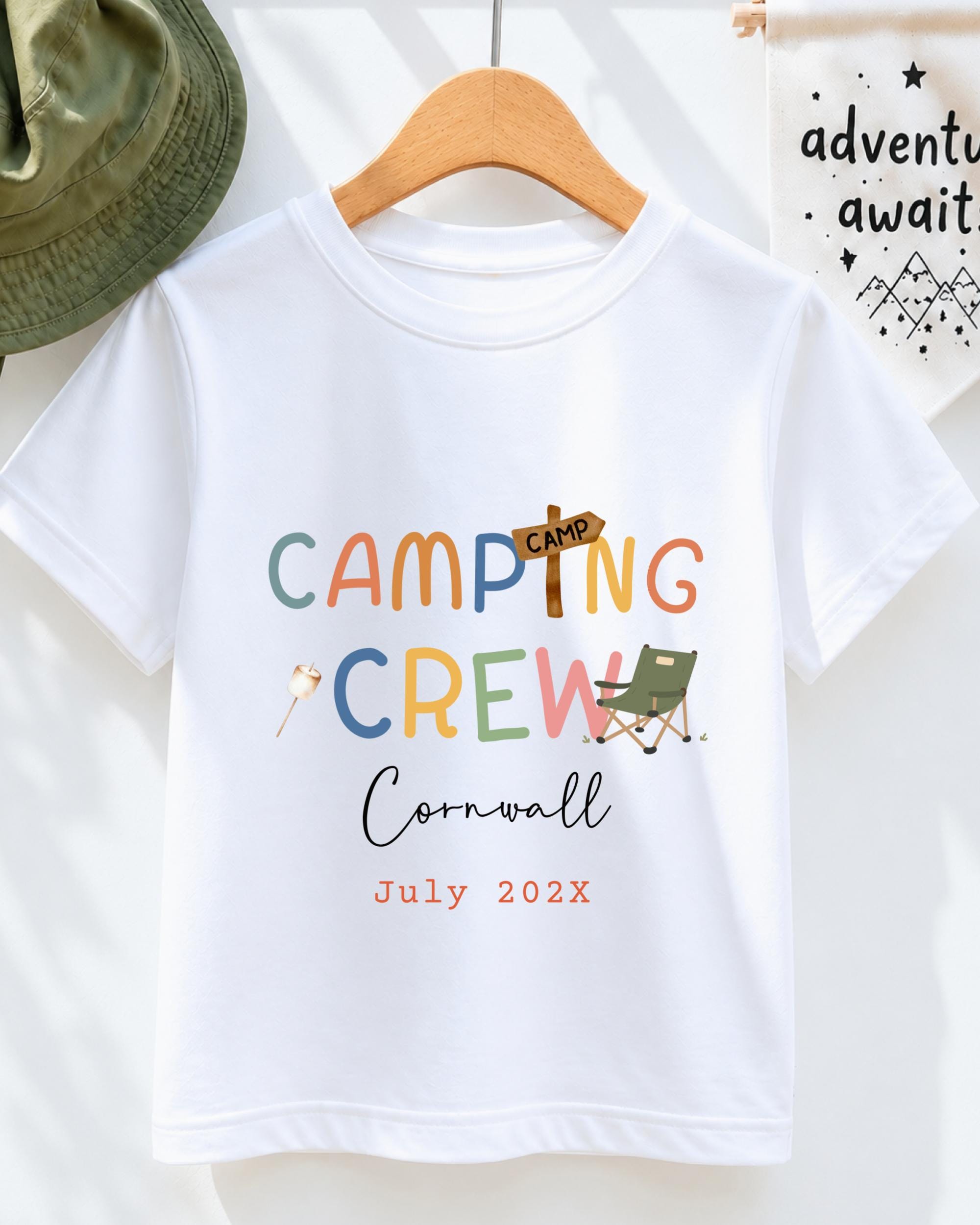 A white t-shirt with colorful text and graphics, including the words "Camping Crew" and "Cornwall" along with a camping-related image. The t-shirt is hanging on a wooden hanger, and there is a green hat and a white banner with text in the background.