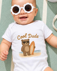 A baby wearing a white onesie with a cartoon bear and a surfboard, smiling and wearing sunglasses.