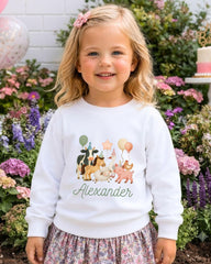 A young girl wearing a white sweater with a cartoon animal design and the name "Alexander" printed on it, standing in front of a garden with colorful flowers.
