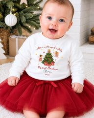 A smiling baby girl wearing a white sweater with a Christmas tree design and a red tulle skirt, sitting on a white carpet in front of a Christmas tree.